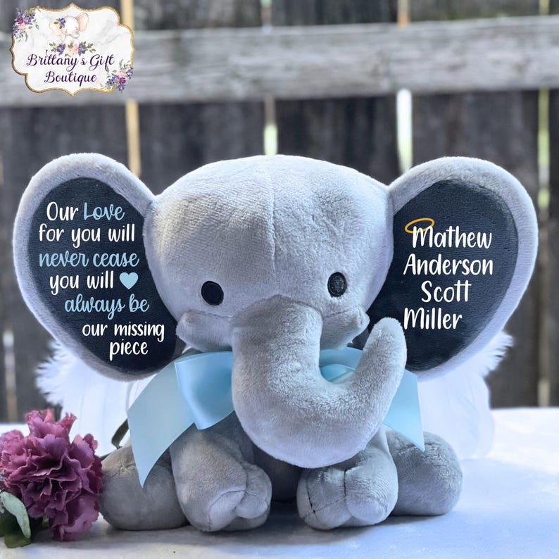 Może przedstawiać: Szary pluszowy słoń z niebieskimi skrzydłami i czarną filcową metką na ucho. Metka na ucho zawiera tekst "Our Love for you will never cease you will always be our missing piece" po jednej stronie i "Mathew Anderson Scott Miller" po drugiej.