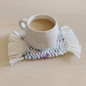 Tiny rug looking coaster with off-white fringes. Main body changes randomly between white, purple, and teal. Moss knit pattern. Photo shows a mug on it.