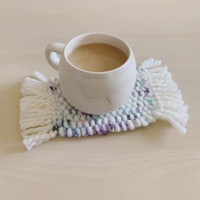 Tiny rug looking coaster with off-white fringes. Main body changes randomly between white, purple, and teal. Moss knit pattern. Photo shows a mug on it.