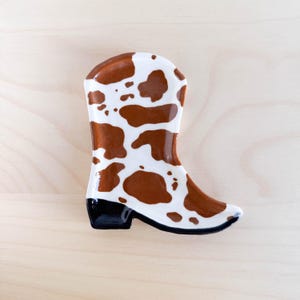 May include: A ceramic boot-shaped dish with a brown and white cow print design. The dish has a black rim around the bottom.