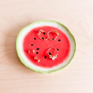 Pode incluir: Um prato de cerâmica em forma de melancia com uma borda verde contém três anéis de ouro e dois brincos de ouro. Os anéis e os brincos estão dispostos no desenho de melancia vermelho e preto.