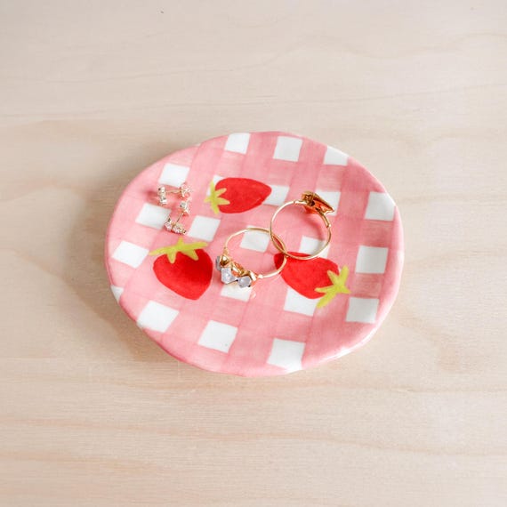 A hand-built gingham strawberry ceramic dish that's so specific and charming it stops your scroll — 62 favorites on a $104 piece proves people genuinely want it.