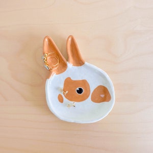 Pode incluir: Um prato de cerâmica branca em forma de coelho com manchas marrons e um olho preto. O prato é decorado com anéis e brincos dourados.