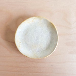 May include: A small, white ceramic dish with a slightly irregular, rounded shape and a light yellow rim. The dish has a subtle, crackled glaze.