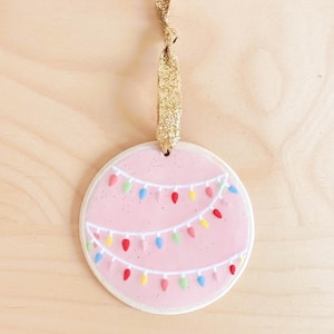 Op de afbeelding: Een ronde, roze kerstversiering met een witte rand en een gouden glitterlint. De versiering heeft twee rijen kleurrijke kerstverlichting. De lampjes zijn rood, geel, blauw en groen.