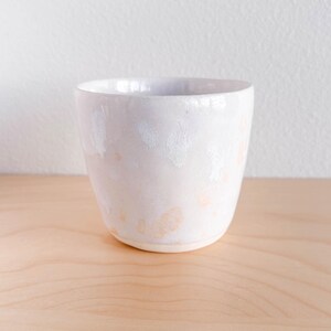 May include: A white ceramic cup with a speckled glaze. The cup has a rounded base and a slightly flared rim. The glaze has a soft, pearlescent sheen with subtle peach-colored spots.