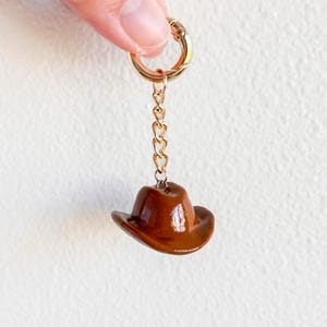 Op de afbeelding: Een miniatuur bruine cowboyhoed bedel, opgehangen aan een goudkleurige ketting en sluiting. De hoed heeft een glanzende afwerking en is bevestigd aan een kleine gouden ketting. De sluiting wordt door vingers vastgehouden.
