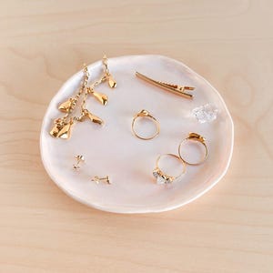 May include: A collection of gold-toned jewelry displayed on a small, light pink ceramic dish. The jewelry includes earrings, rings, and a hair clip. The earrings have a dangling design, and the rings feature small, clear stones. The dish sits on a light wood surface.