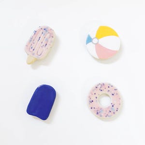 May include: Four ceramic decorative pieces including a light pink popsicle with blue speckles, a round beach ball with yellow, white, and pink segments, a solid dark blue popsicle shape, and a light pink ring-shaped piece with blue speckles.