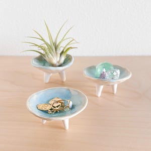 Op de afbeelding: Drie kleine, lichtblauwe keramische schalen met elk drie witte poten. De schalen staan op een licht houten oppervlak. Eén schaal heeft een klein luchtplantje, een andere heeft een groene kristal en de derde heeft gouden ringen.