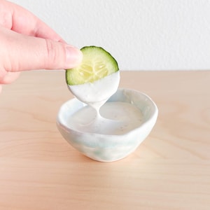 May include: A white ceramic bowl filled with a creamy white dip. A slice of cucumber is being dipped into the dip.
