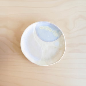 Pode incluir: Um pequeno prato de cerâmica branca com um esmalte de cor púrpura claro e amarelo. O prato tem uma forma irregular e uma borda ligeiramente elevada.