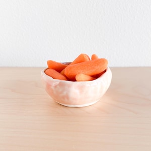 May include: A small, light pink ceramic bowl filled with bright orange baby carrots. The bowl has a slightly irregular shape and a textured surface. The carrots are piled high, creating a visually appealing arrangement.