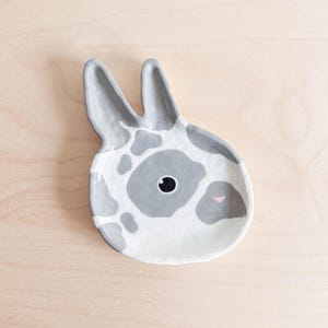 Pode incluir: Um prato de cerâmica em forma de cabeça de coelho. O prato é branco com manchas cinzas e um nariz rosa. Os olhos do coelho são pretos.