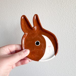 May include: A brown and white ceramic dish shaped like a rabbit's face. The dish has a white rim and a brown interior. The rabbit's face has a black eye and a pink nose.
