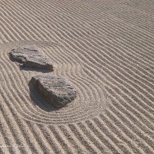 K&ouml;nnte beinhalten: Ein Zen-Garten mit geharktem Sand und zwei grauen Steinen. Der Sand ist mit geschwungenen und geraden Linien gemustert, was ein Gef&uuml;hl von Frieden und Ruhe erzeugt. Die Steine sind rau und strukturiert.