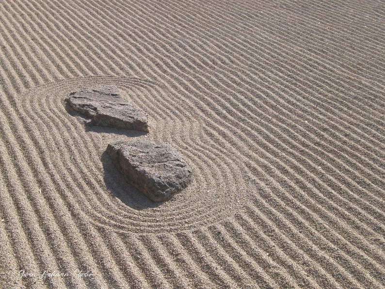 K&ouml;nnte beinhalten: Ein Zen-Garten mit geharktem Sand und zwei grauen Steinen. Der Sand ist mit geschwungenen und geraden Linien gemustert, was ein Gef&uuml;hl von Frieden und Ruhe erzeugt. Die Steine sind rau und strukturiert.