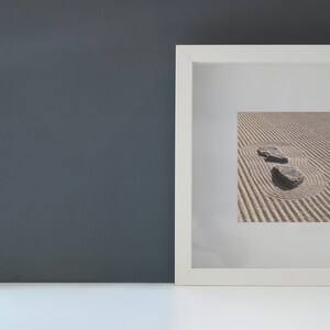 K&ouml;nnte beinhalten: Ein wei&szlig; gerahmter Druck mit einem Zen-Garten mit zwei Steinen in einem Sandmuster. Rechts vom Rahmen befindet sich ein kleiner Kaktus in einem wei&szlig;en Topf. Der Hintergrund ist eine dunkelgraue Wand.