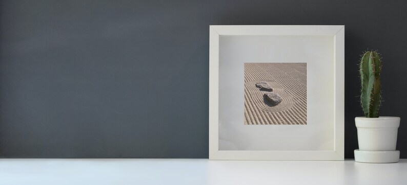 K&ouml;nnte beinhalten: Ein wei&szlig; gerahmter Druck mit einem Zen-Garten mit zwei Steinen in einem Sandmuster. Rechts vom Rahmen befindet sich ein kleiner Kaktus in einem wei&szlig;en Topf. Der Hintergrund ist eine dunkelgraue Wand.