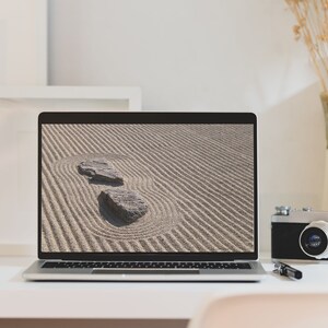 K&ouml;nnte beinhalten: Ein moderner Arbeitsplatz mit einem Laptop, der ein Zen-Garten-Bild mit zwei Steinen zeigt. Eine Vintage-Kamera, Bleistifte, eine Kaffeetasse und dekorative Gegenst&auml;nde befinden sich ebenfalls auf dem wei&szlig;en Schreibtisch. Die Szene vermittelt Ruhe und Kreativit&auml;t.