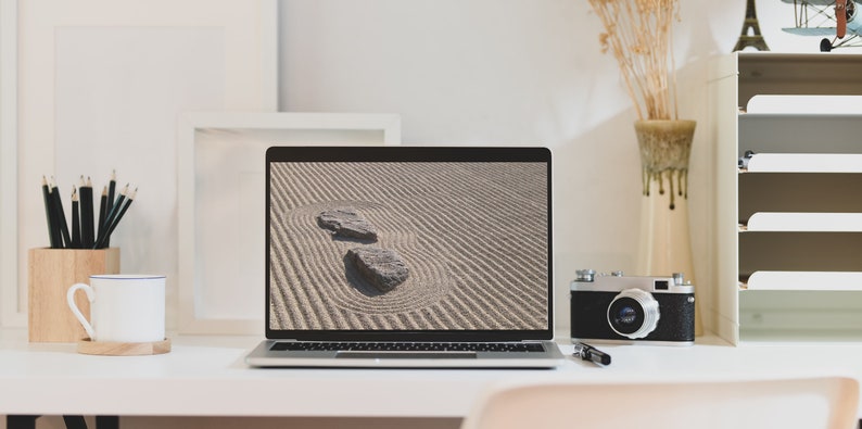 K&ouml;nnte beinhalten: Ein moderner Arbeitsplatz mit einem Laptop, der ein Zen-Garten-Bild mit zwei Steinen zeigt. Eine Vintage-Kamera, Bleistifte, eine Kaffeetasse und dekorative Gegenst&auml;nde befinden sich ebenfalls auf dem wei&szlig;en Schreibtisch. Die Szene vermittelt Ruhe und Kreativit&auml;t.