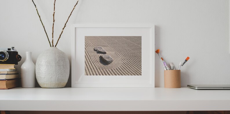 K&ouml;nnte beinhalten: Ein wei&szlig; gerahmter Druck eines Zen-Gartens mit zwei Steinen in einem Sandmuster. Der Druck befindet sich auf einer wei&szlig;en Oberfl&auml;che mit einer Vase, einer Kamera, B&uuml;chern und Kunstzubeh&ouml;r.