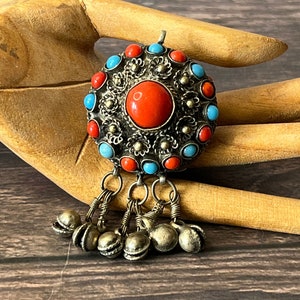 May include: A silver pendant with a round, turquoise and red stone design. The pendant has a silver filigree design and is accented with small silver bells.