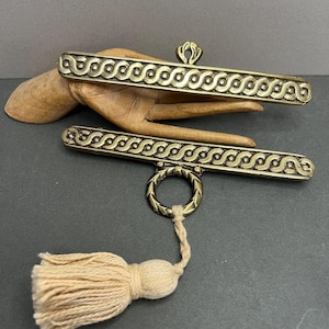 May include: Two antique brass toggle clasps with a decorative wave pattern and a cream-coloured tassel. The clasps are displayed on a wooden hand against a dark grey background. The toggle clasp is a jewellery finding.