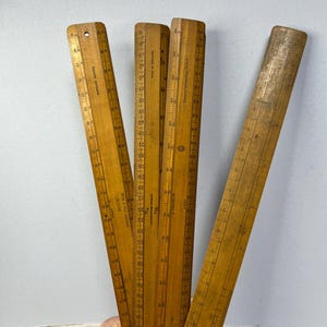 May include: Four vintage wooden rulers with markings in inches. The rulers are all slightly different lengths and have different brands printed on them. The rulers are made of a light brown wood and have a slightly worn look.