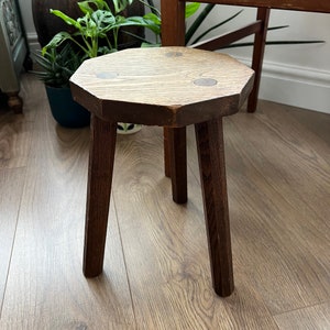 Op de afbeelding: Een kleine, houten kruk met een zeshoekige bovenkant en drie poten. De kruk is gemaakt van donkerbruin hout en heeft een rustieke uitstraling.