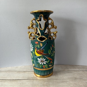 Könnte beinhalten: Eine dunkelgrüne Keramikvase mit goldenen Akzenten und einem Vogel- und Blumenmuster. Die Vase hat eine einzigartige Form mit ausgeschnittenen Griffen und einem goldenen Rand. Der Vogel ist gelb, rot und braun, und die Blumen sind weiß.