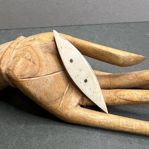 Könnte beinhalten: Ein cremefarbenes, längliches Objekt mit zwei kleinen, dunklen Punkten. Das Objekt hat ein spitzes Ende und liegt auf einer geschnitzten Holzhand. Die Hand ist hellbraun und hat detaillierte Schnitzereien.