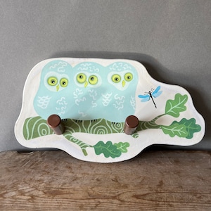 May include: A white wooden owl coat rack with three owls and a dragonfly. The owls have green eyes and are perched on a branch with green leaves. The rack has two brown wooden hooks.
