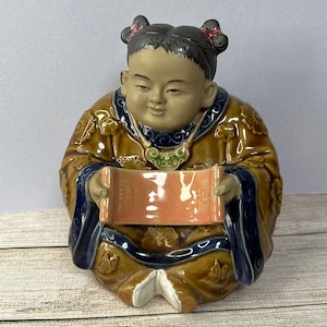 May include: A ceramic figurine of a child in traditional Chinese clothing, sitting with a scroll in their hands. The figurine is painted in shades of brown, blue, green, and pink.