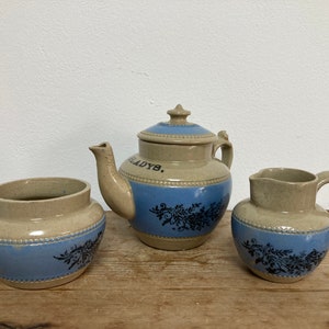 May include: A vintage tea set comprising a teapot, creamer and sugar bowl. The set is made of stoneware and features a blue and white floral design. The teapot has the words "Old Ladys" written on it.