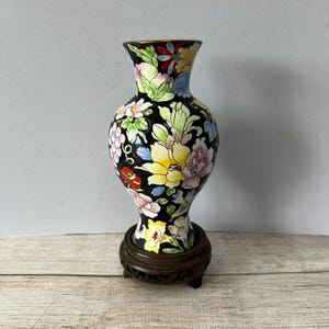 May include: A small black vase with a floral design in shades of pink, yellow, blue, and red. The vase is on a wooden stand with three legs.