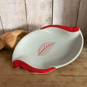 May include: White ceramic dish with a red rim and a red leaf design. The dish is slightly curved and has a rounded shape.