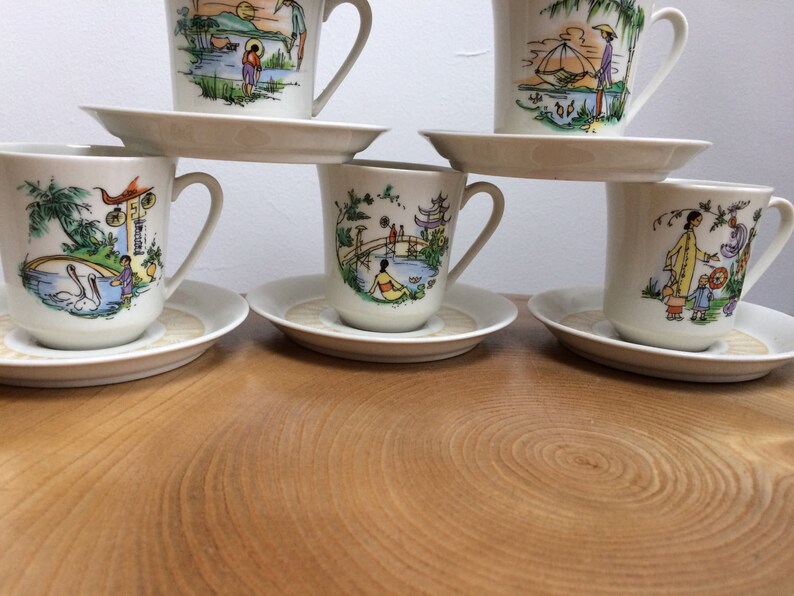 May include: Set of six white teacups and saucers with colorful Asian-inspired designs. Each cup features a different scene with people, buildings, and nature.