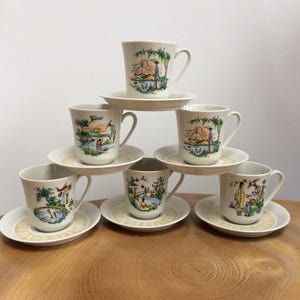 May include: Set of six white porcelain teacups and saucers with Asian-inspired designs. Each cup and saucer features a different scene with people, boats, and buildings.