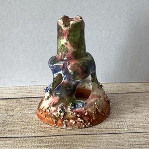 May include: A ceramic candle holder with a unique, abstract design. The holder is painted in shades of green, blue, pink, and brown, and has a textured surface. The holder has a hole in the center for a candle.