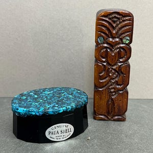 May include: A small black oval box with a lid inlaid with blue and green crushed paua shell, featuring a white label that reads GENUINE PAUA SHELL MADE BY ARIKI NEW ZEALAND, alongside a carved brown wooden tiki figure with inlaid shell eyes.