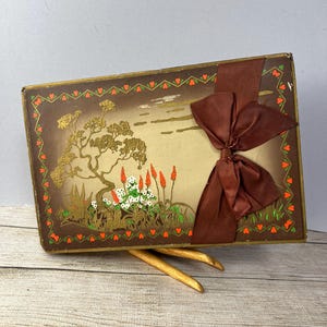 May include: A vintage gift box with a brown background and a gold and green border. The box features a painted scene of a tree, flowers, and a lake. A brown satin ribbon is tied around the box with a bow.