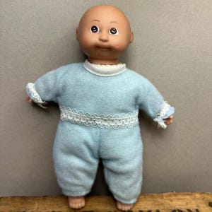 Könnte beinhalten: Eine kahle Babypuppe mit heller Haut, braunen Augen und einem kleinen Lächeln. Die Puppe trägt einen hellblauen langärmeligen Strampler mit weißer Spitzenborte am Kragen, an der Taille und an den Ärmelbündchen, der eine weiche Textur aufweist.