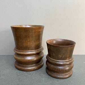 May include: Two turned wooden vessels with a dark brown finish. The larger vessel has a flared top and a base with three rounded sections. The smaller vessel has a similar design. Both are set against a neutral background.