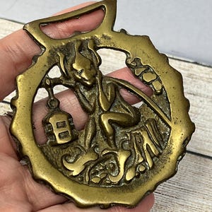 Könnte beinhalten: Ein Messing-Pferdeanhänger mit einem detaillierten Design einer Figur, die eine Laterne hält. Die Figur sitzt auf einer Bank mit der Aufschrift "Spanien" darunter.