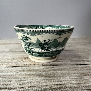 Puede incluir: Un cuenco de cerámica blanco y verde con una escena de paisaje detallada. El cuenco presenta un borde verde oscuro y una base de pedestal. El diseño del paisaje incluye una persona a caballo, árboles y montañas.