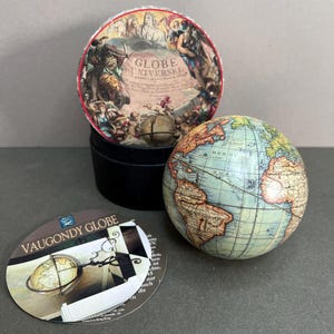 以下が含まれることがあります： 詳細な世界地図が描かれたミニチュア地球儀。黒い箱と丸いタグの隣にあります。地球儀の直径は約7.6cmです。箱の蓋には「GLOBE UNIVERSEL」の文字が入った歴史的なイラストが描かれています。