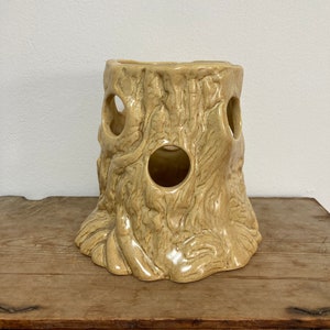 May include: A ceramic planter shaped like a tree stump with three holes cut out. The planter is a light beige colour and has a textured surface.