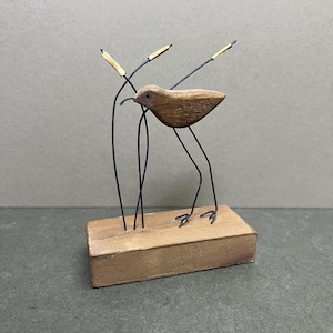 May include: A wooden bird sculpture with wire legs and a beak, perched on a rectangular wooden base. The bird has a brown body and is accompanied by three stalks with gold accents. The sculpture is set against a gray background.