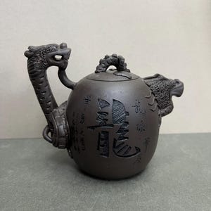 May include: Dark brown ceramic teapot with dragon-shaped handle and spout. The teapot has a rounded body with Chinese characters etched on the surface. The handle and spout are detailed with dragon scales and features.